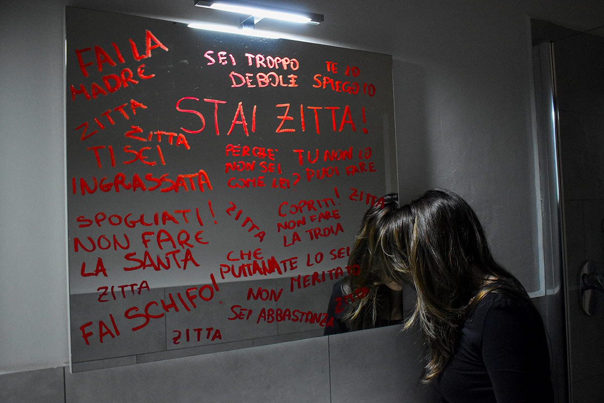 donna davanti a uno specchio che riporta una serie di frasi maschiliste, tipo: "Stai zitta!"